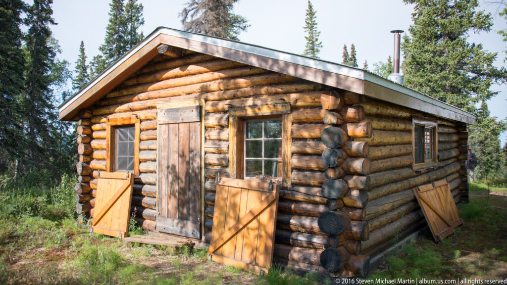 Small log cabin with open window shutters, plank door, and metal stovepipe.