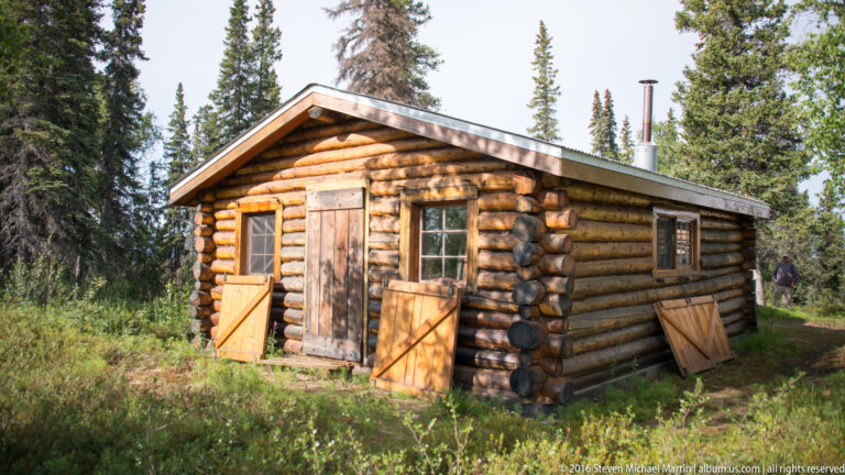 Log cabin with open shutters stands in a grassy clearing among trees.