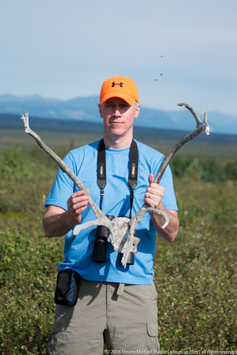 Man in an orange cap holds a large shed antler with binoculars around his neck.