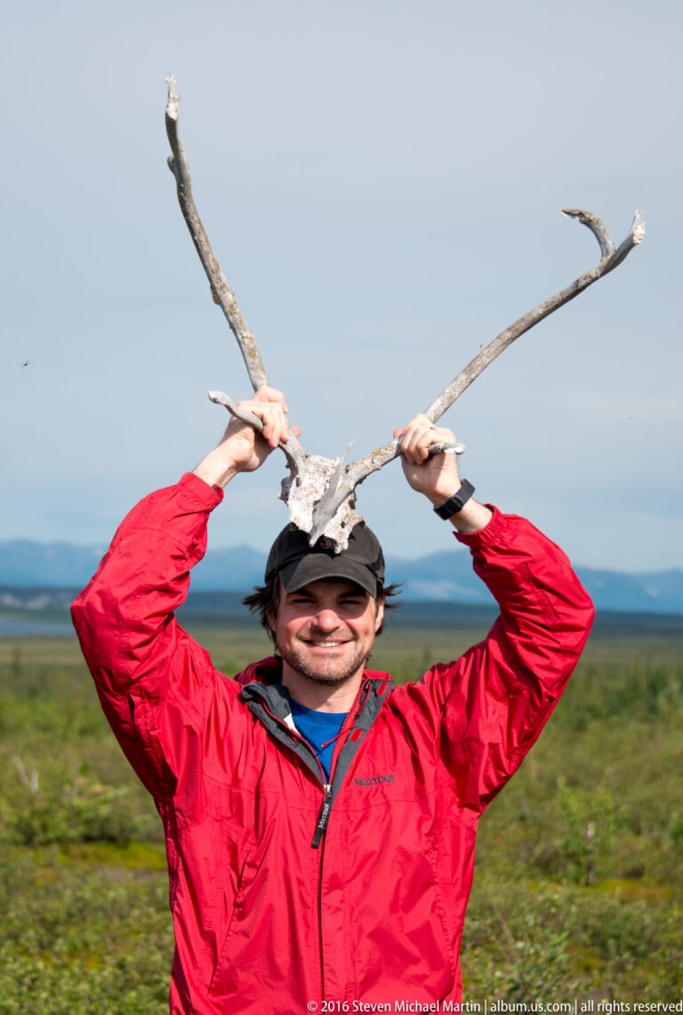 Man in a red jacket holds a large branching antler above his head.