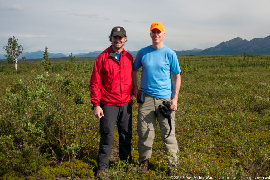 Two visitors in red and blue outdoor clothing stand together on tundra.