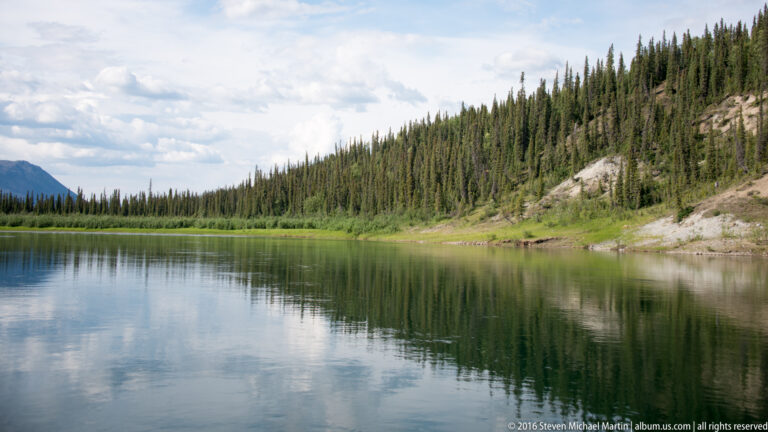 A spruce-covered slope and pale sandy shore reflect in a calm river beneath clouds.