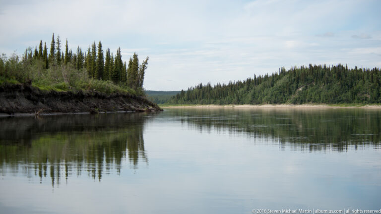 A broad river curves between forested banks, with an undercut near shore and a pale sandbar farther away.