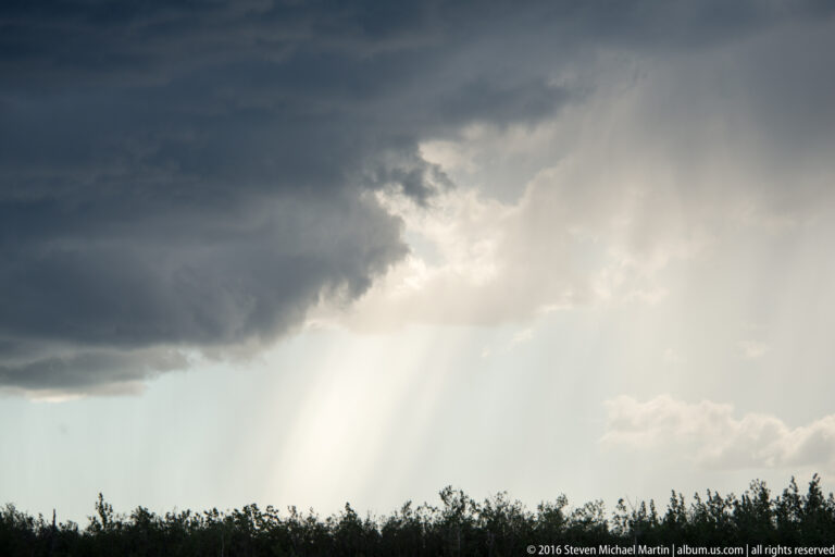 Sun rays stream beneath a dark cloud above a narrow line of trees.