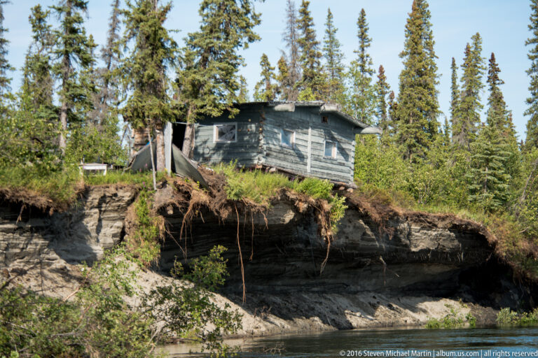 A weathered house stands above a steep eroded bank with exposed soil and dangling roots.