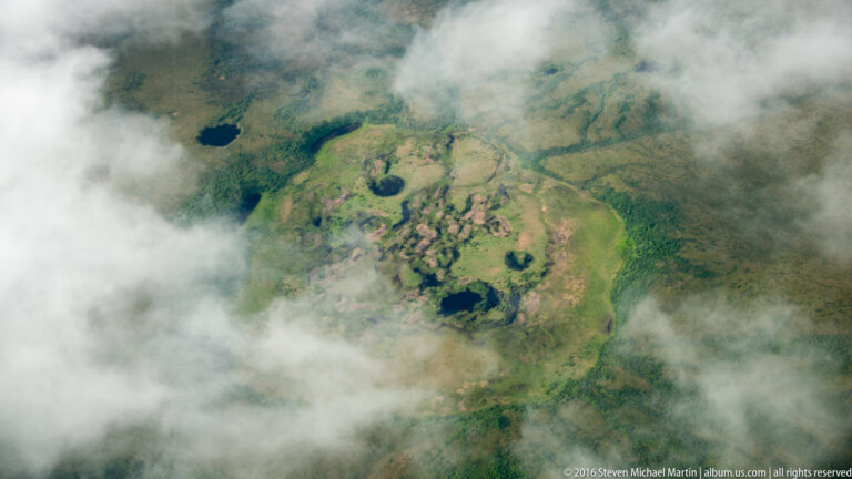 Broken clouds reveal an aerial view of a green tundra basin dotted with dark ponds and small lakes.