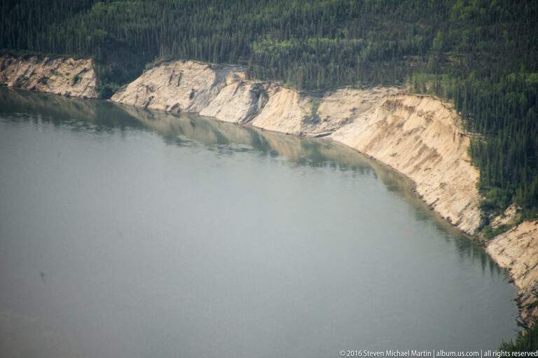 Pale exposed bluffs curve along a broad river beneath a dense spruce-covered bank.