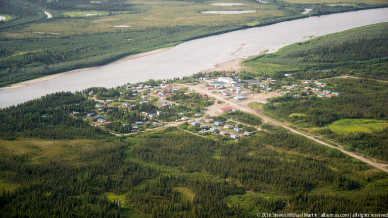 An aerial view shows homes and roads in a forest clearing beside a broad river.