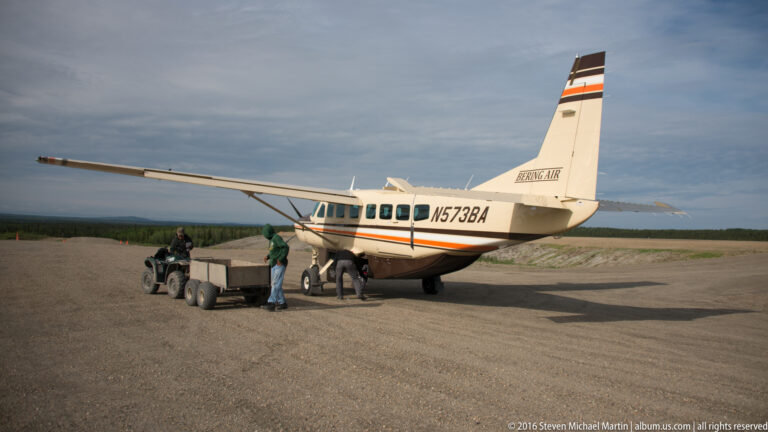 A single-engine high-wing plane is parked on a gravel strip beside a trailer and several people.