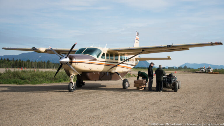 A high-wing airplane faces the camera on a gravel airstrip while people handle equipment beside it.