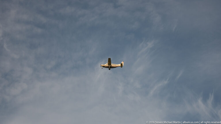 A small single-engine airplane flies in profile across a wide gray-blue sky streaked with thin clouds.