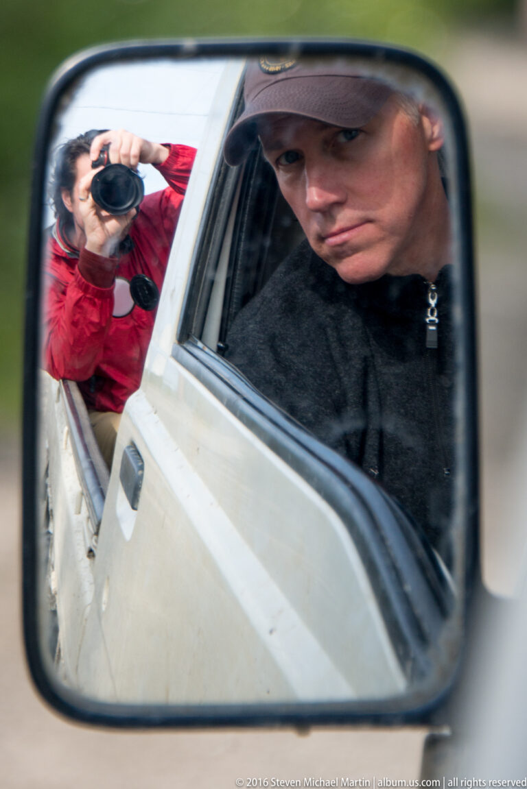 A vehicle side mirror reflects a man inside and a person in a red jacket photographing from outside.