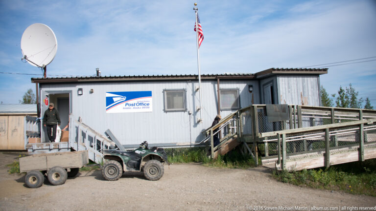 A small post office with a raised ramp stands beside a gravel yard, trailer, and all-terrain vehicle.