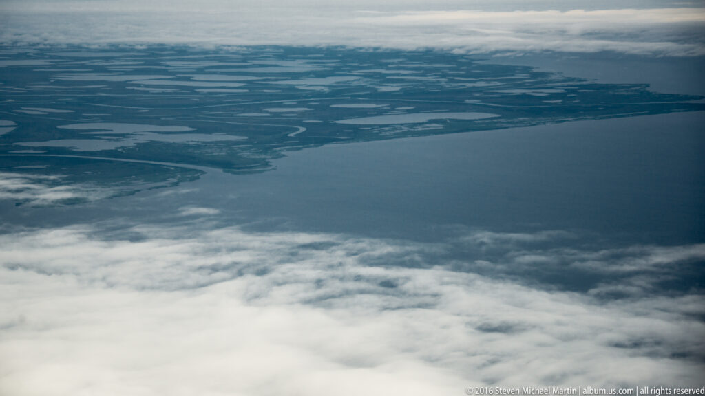 Low clouds cover part of an aerial view of a broad watery plain with distant channels and pools.