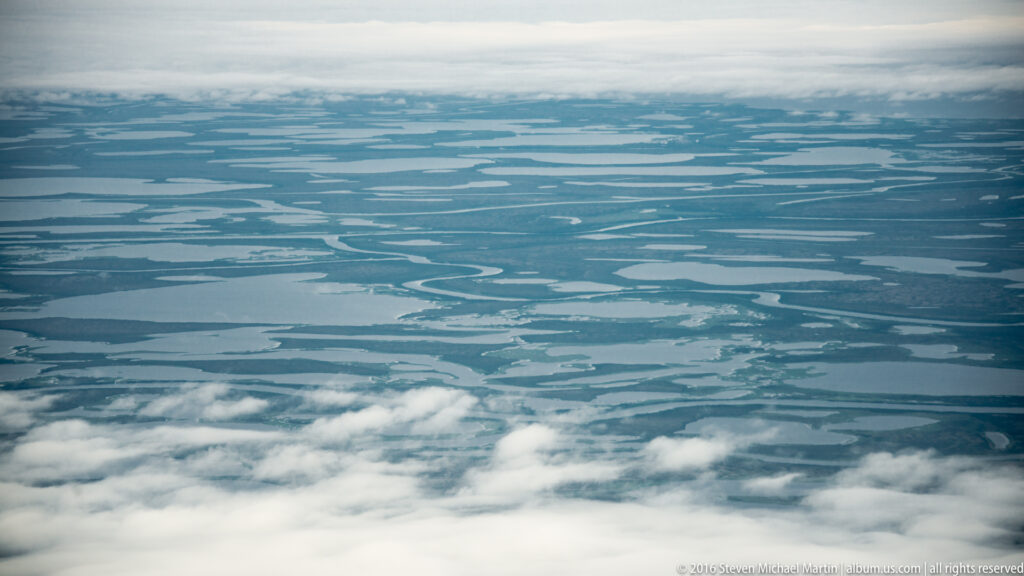 Clouds part above an aerial view of a blue-green plain patterned with ponds and narrow channels.