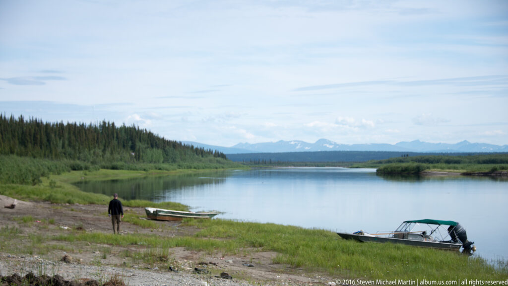 Person stands beside a small boat on a gravel riverbank with another boat at the water’s edge.