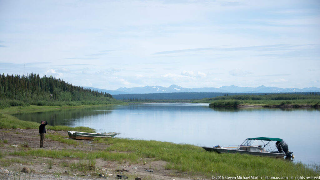 Two small boats rest on a gravel riverbank beside a person and a broad river.