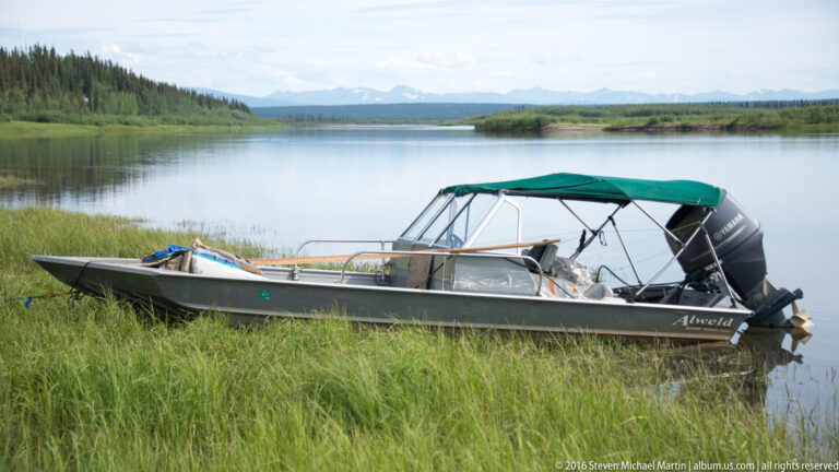 Aluminum motorboat with green canopy and outboard engine rests in grass beside a river.