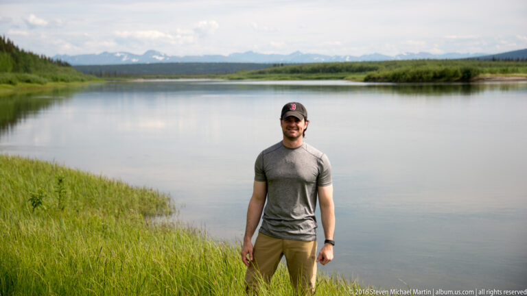 Man in a gray shirt stands on a grassy bank beside a calm river.