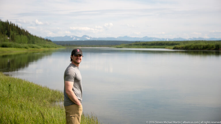 Man in a gray shirt turns toward the camera beside a broad river.