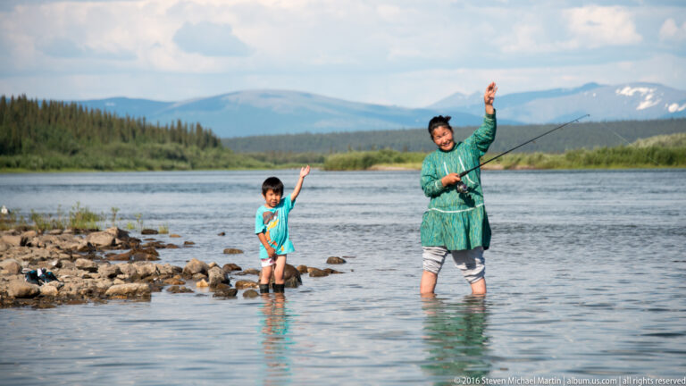 Woman and child stand in shallow river water and wave beside a rocky shore.