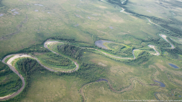 An aerial view shows a wide river looping through open tundra, with forested strips and ponds across the plain.