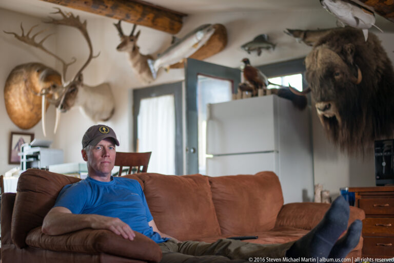 Man sits on a sofa beneath mounted fish, antlers, and a large animal head.