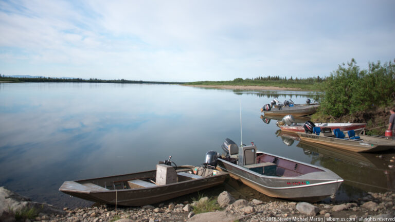 Several small motorboats are moored along a rocky riverbank beneath trees.