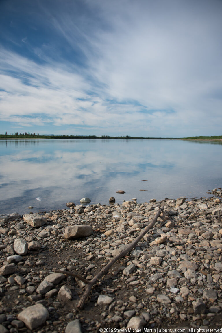 Pebble shoreline and driftwood border a broad river beneath a cloudy sky.