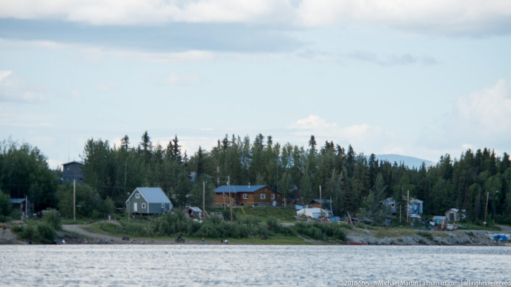 Small homes and utility poles line a wooded village shore across a broad river.