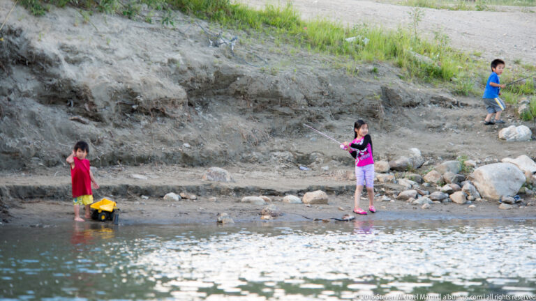 Three children play in shallow water below a rocky, grass-topped riverbank.