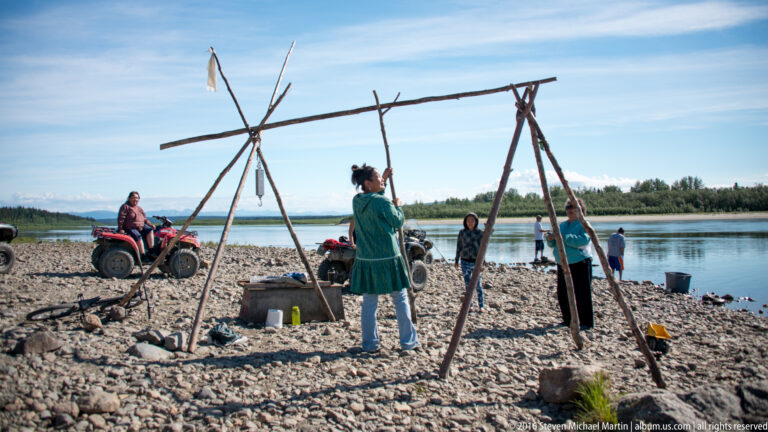 People gather around tall timber frames and equipment on a gravel riverbank.