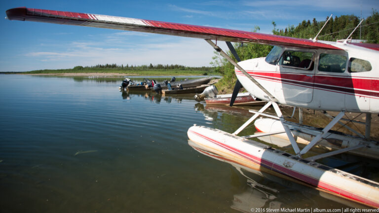 Red-and-white floatplane moored beside small motorboats on a calm river.
