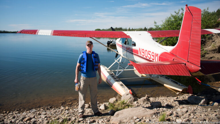Man in a blue life vest stands beside a red-and-white floatplane marked N9059M.