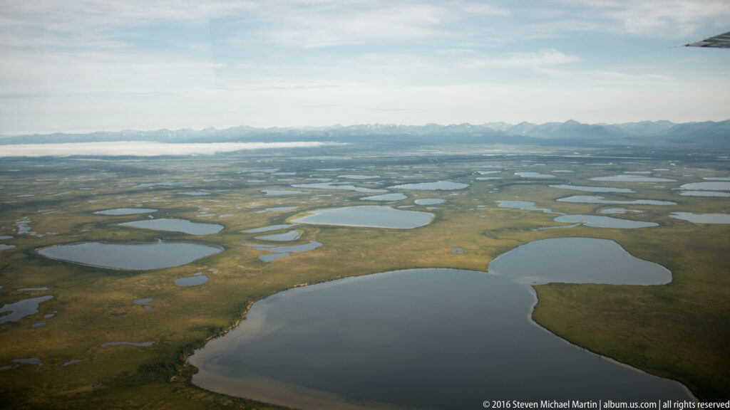 An aerial panorama shows numerous ponds and lakes across open tundra, with distant mountains on the horizon.