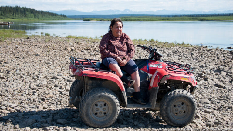 A woman sits on a red ATV on a gravel bar beside a braided river, with distant mountains under a partly cloudy sky.