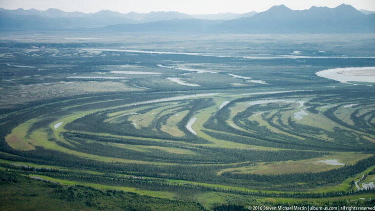 A braided river loops across a green plain beneath distant mountains, with gravel bars dividing its channels.