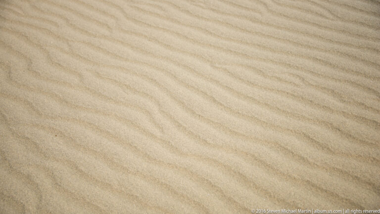 Evenly spaced wind ripples form soft parallel bands across an uninterrupted surface of pale sand.