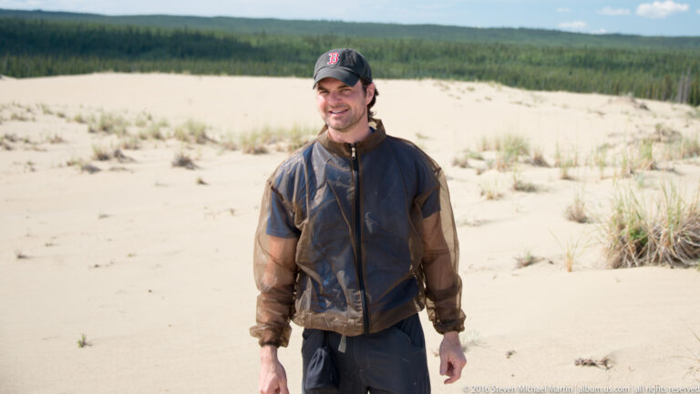A man in a dark cap and outdoor jacket stands on pale sand, with sparse grasses and spruce forest behind.