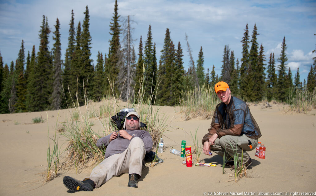 One person reclines and another sits beside dune grass, with bottles and small supplies on the sand.
