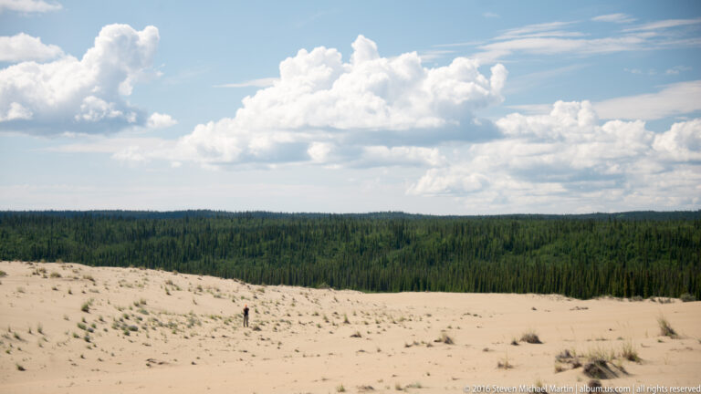 A tiny person stands on a wide pale dune below a dark forest line and towering white clouds.