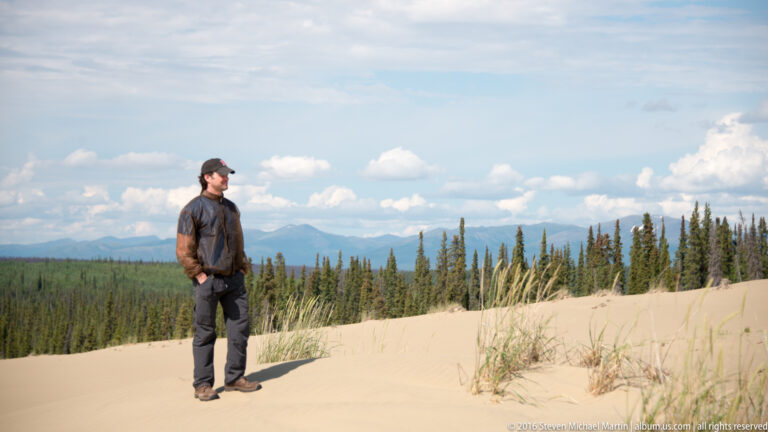 A visitor stands among dune grasses, with forest and distant mountains beneath a bright sky.