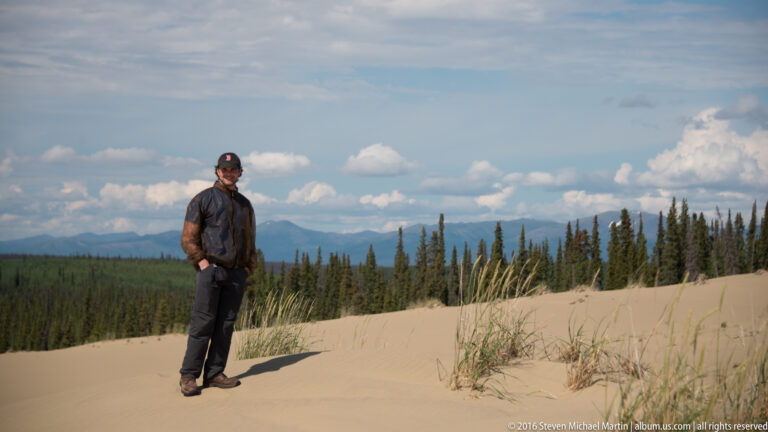 A man stands left of center on a sandy ridge, casting a long shadow before forest and mountains.