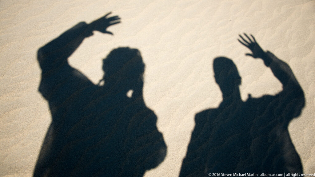 Two sharply defined human shadows, both with raised arms, stretch across pale, lightly rippled dune sand.
