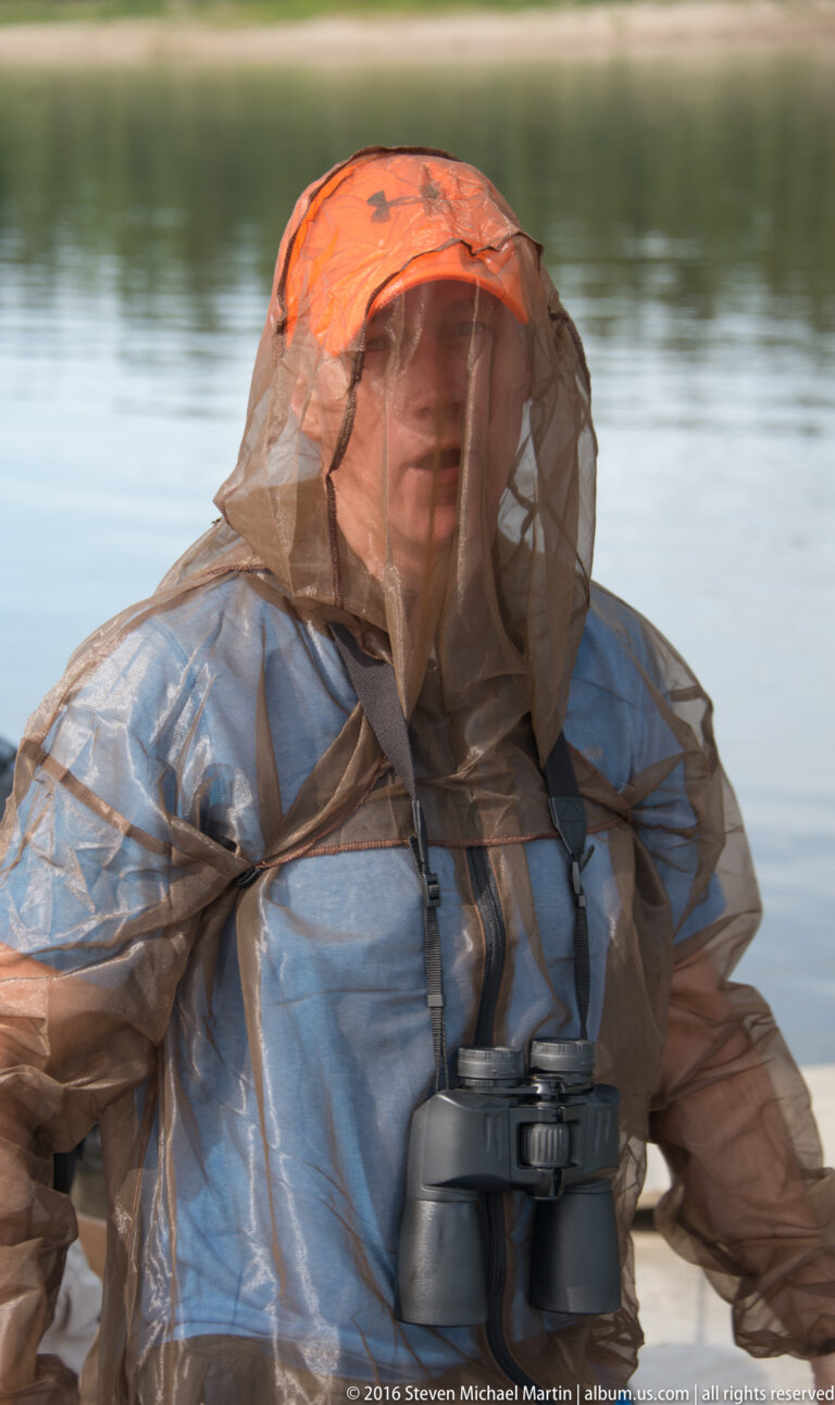 A person in an orange-topped mesh head net and outdoor jacket wears binoculars beside softly blurred water.