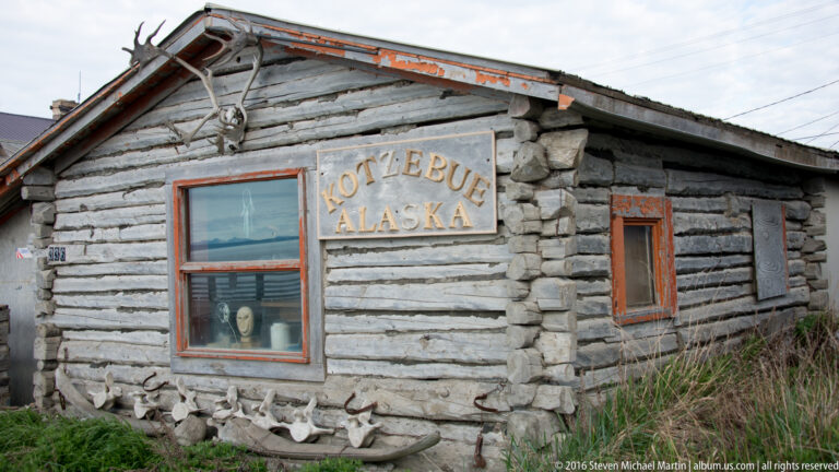 A weathered log building bears a Kotzebue Alaska sign between orange-trimmed windows, with grass at its base.