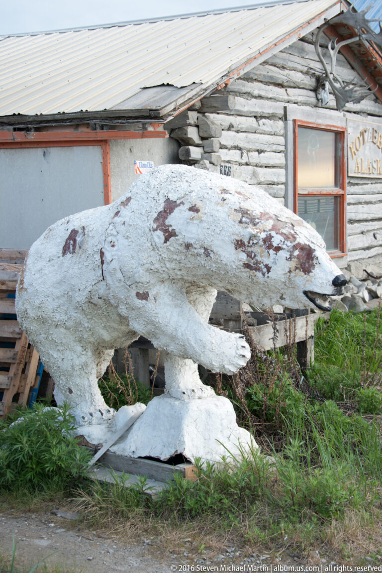 A large pale animal-shaped sculpture stands on supports beside a weathered log building and grass.