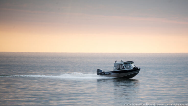 A small covered motorboat speeds across calm water, trailing spray beneath a pale evening sky.