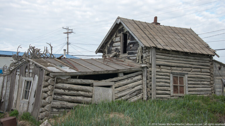 A weathered log house with damaged roof sections has many antlers arranged along the eaves.