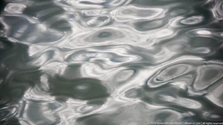 Silvery cloud reflections form distorted, flowing shapes across dark rippled water, filling the entire close-up frame.